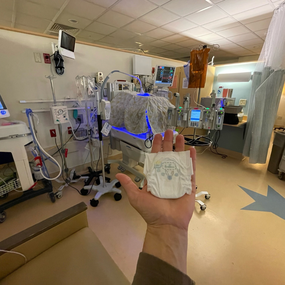 Hand holding preemie diaper in NICU room.